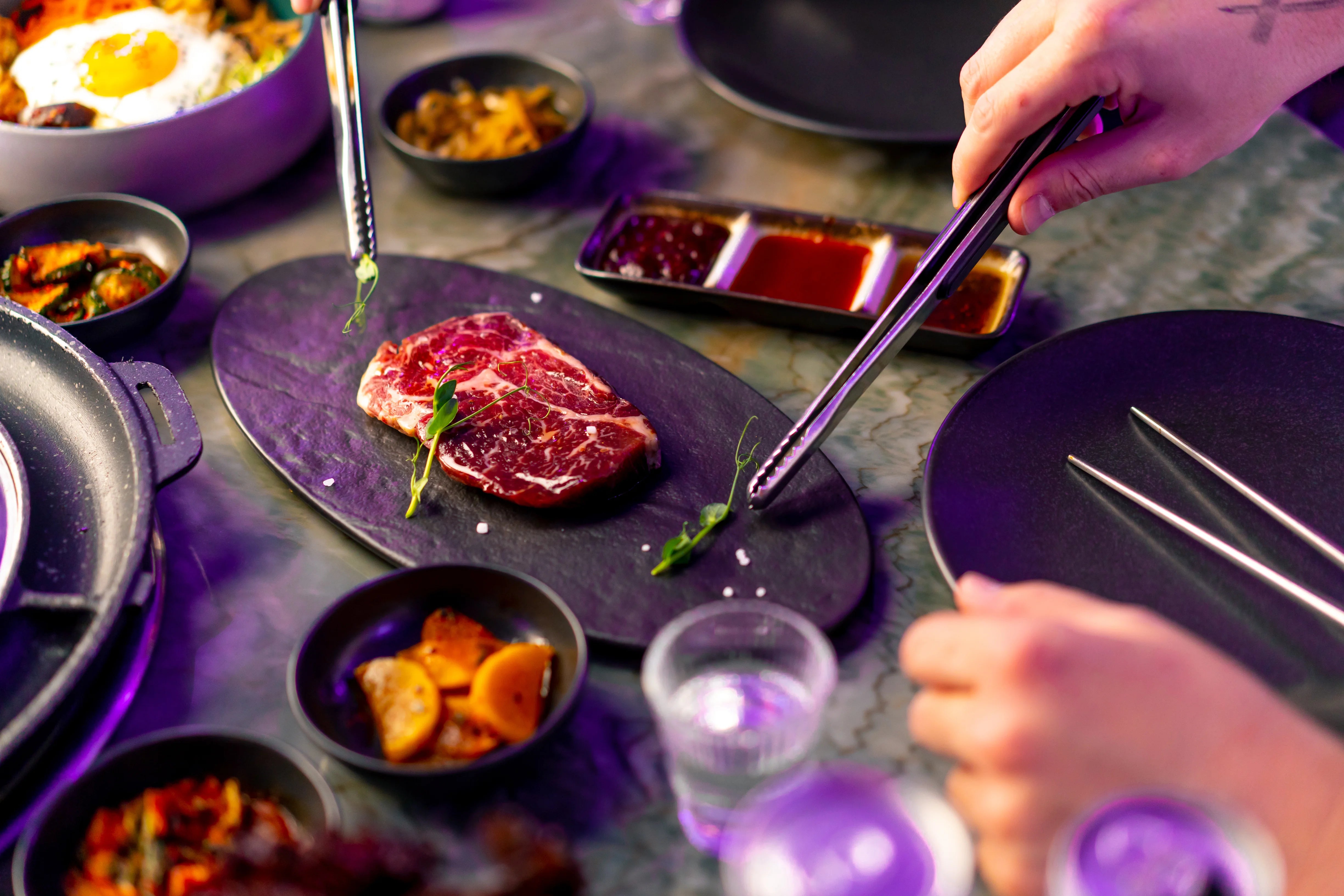 Man sieht ein sehr schön marmoriertes Stück Dry-Aged-Fleisch, umringt von asiatischen Saucen und Gerichten