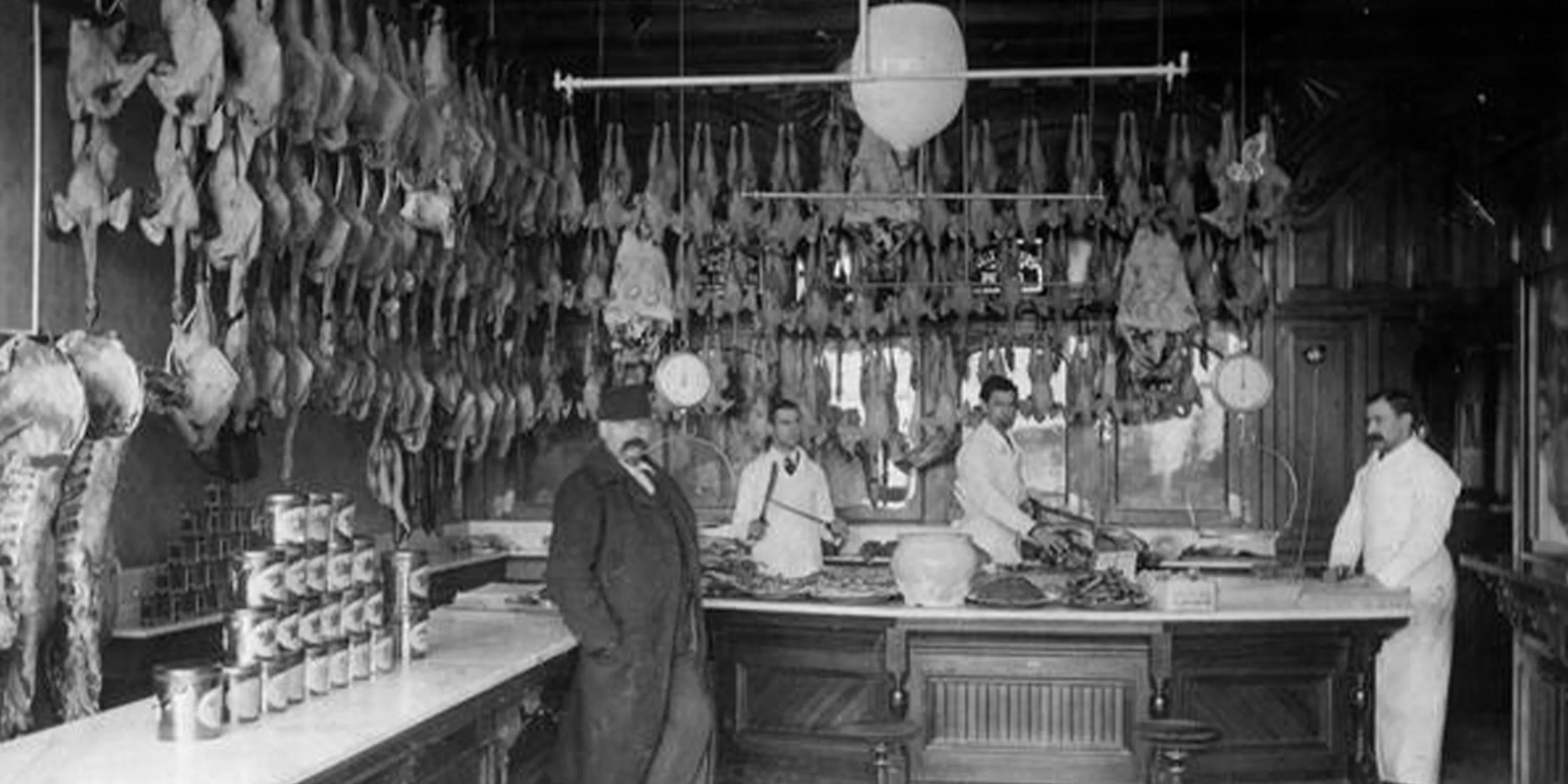 Man sieht eine alte Fleischerei mit drei Fleischern und einem Kunden, in der viel Fleisch zum Trocknen und zur Ausstellung hängt.