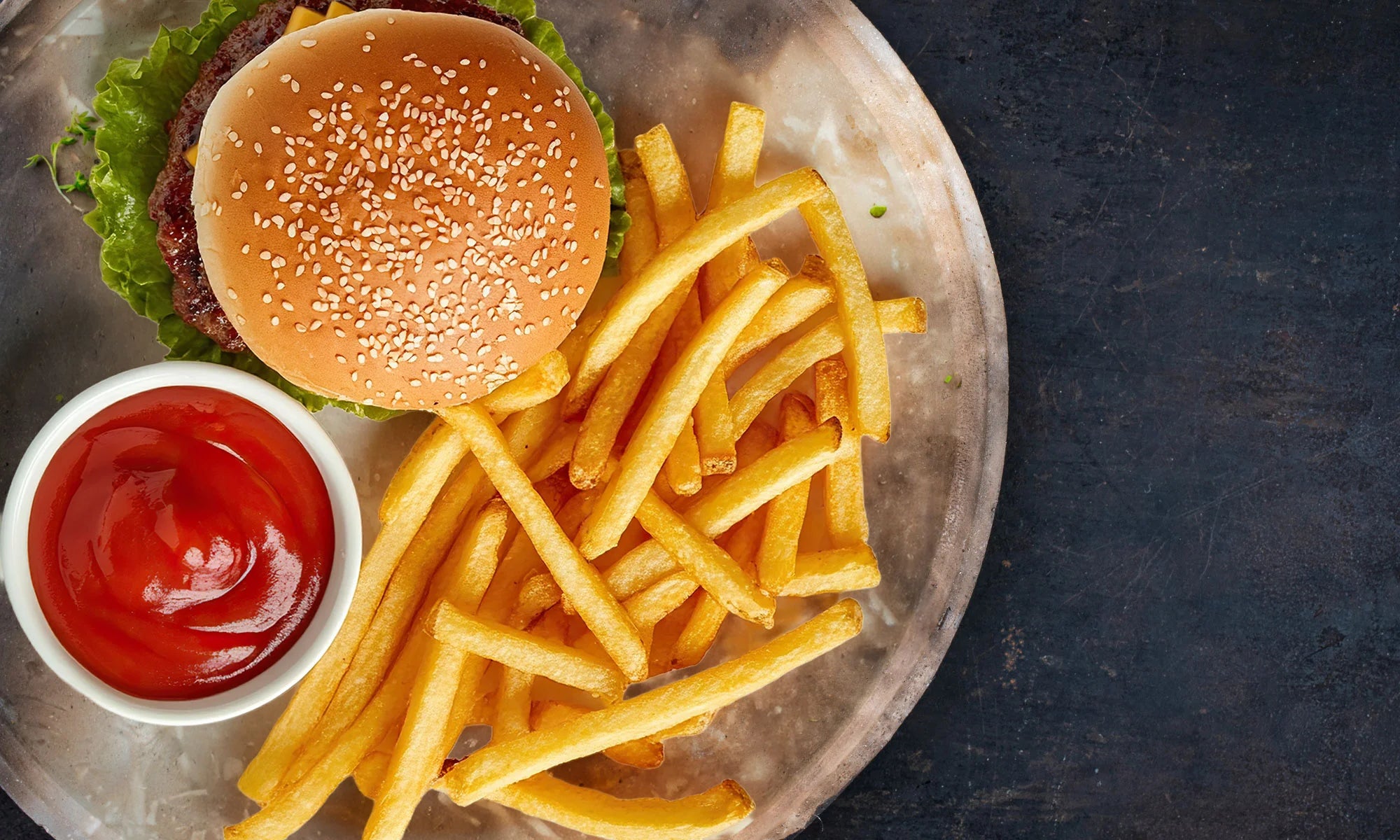 Man sieht einen Teller mit Ketchup, einem ungesunden Hamburger und Pommes